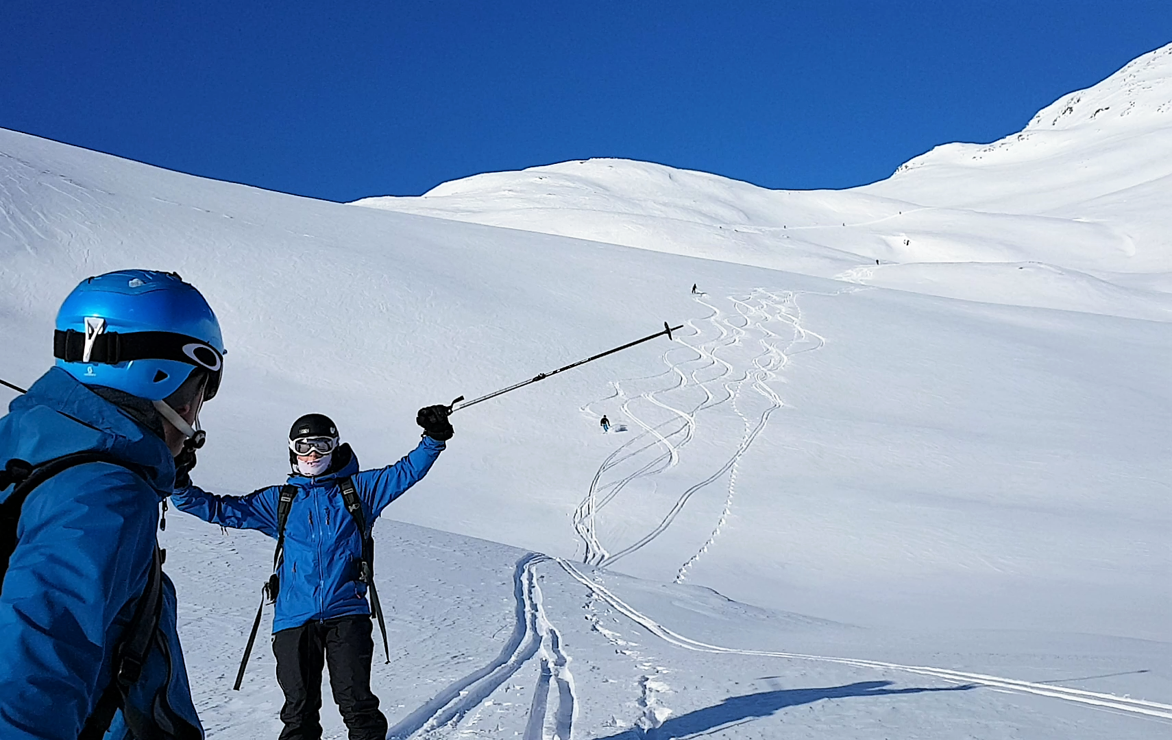 To personar i blått stend på ein snøvkledd fjelltopp, ein viftar med skistaven.