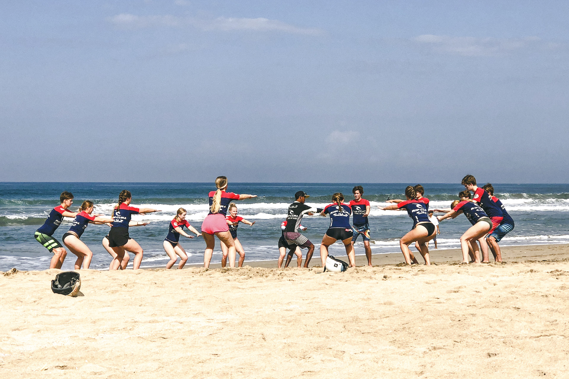 Ei gruppe elevar stend i sirkel mot kvarandre og strekk seg i yoga posisjon med bøygde kne og ustrakte armar på stranda.